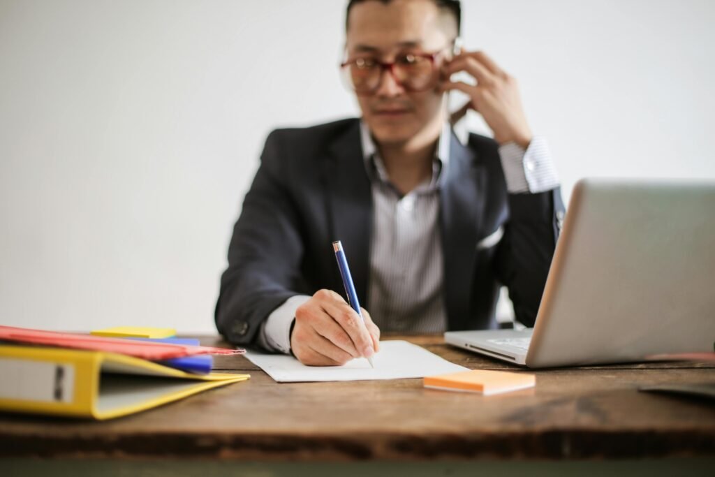 Businessman talking on phone while writing notes and using a laptop at desk.